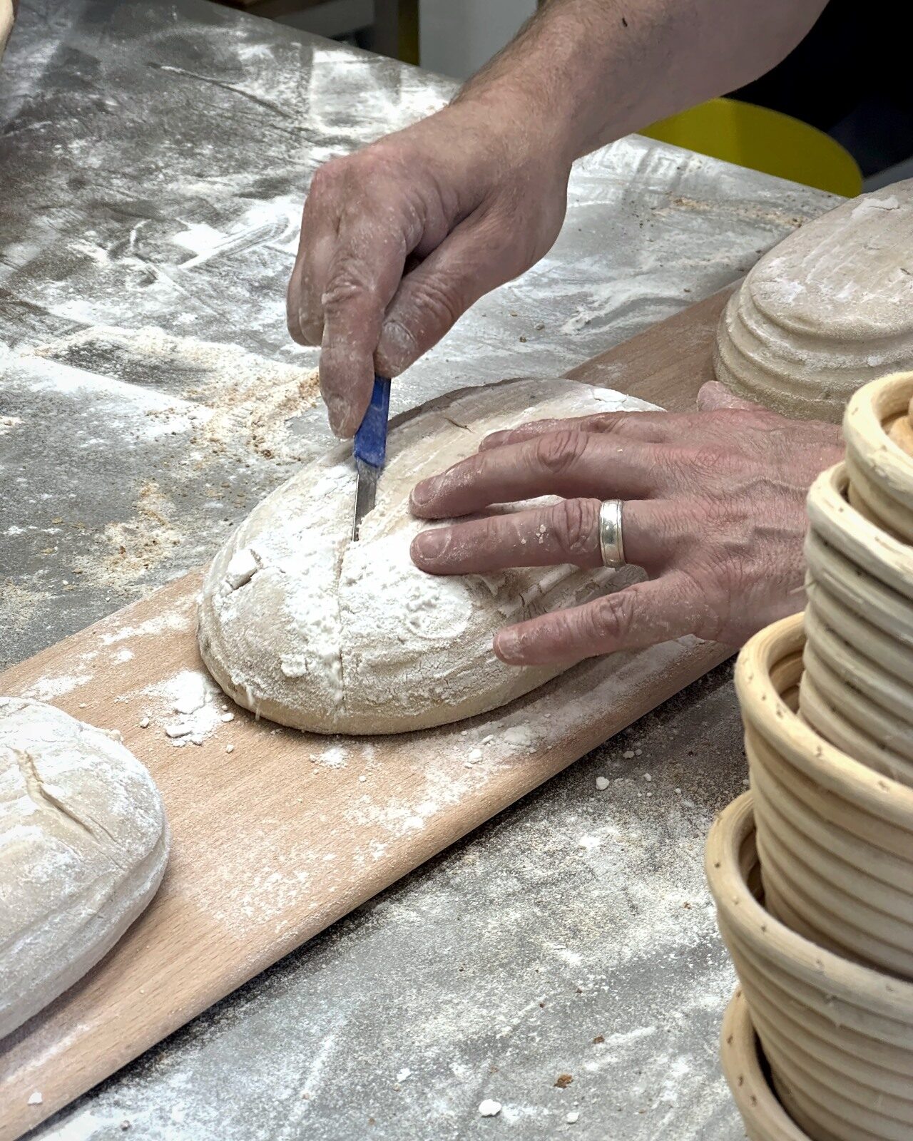 Hands scoring a sourdough loaf with a lame, proving baskets alongside, at The Globe Bakery